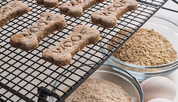 Homemade Dog Treats Bone shaped dog treats cool on a wire rack next to bowls of oats, flour, and eggs.
