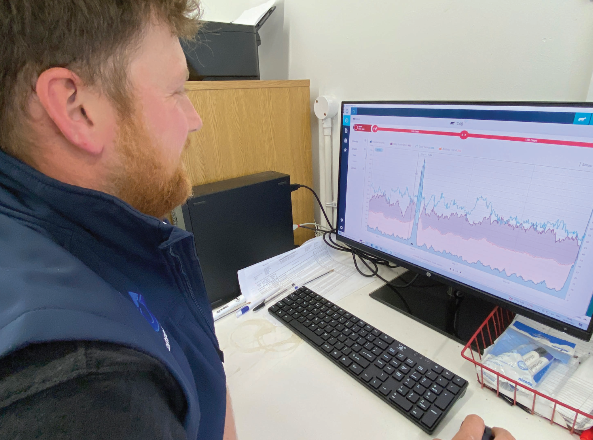 A close-up of a farmer looking at a computer monitor displaying colorful health and activity graphs for a dairy herd.