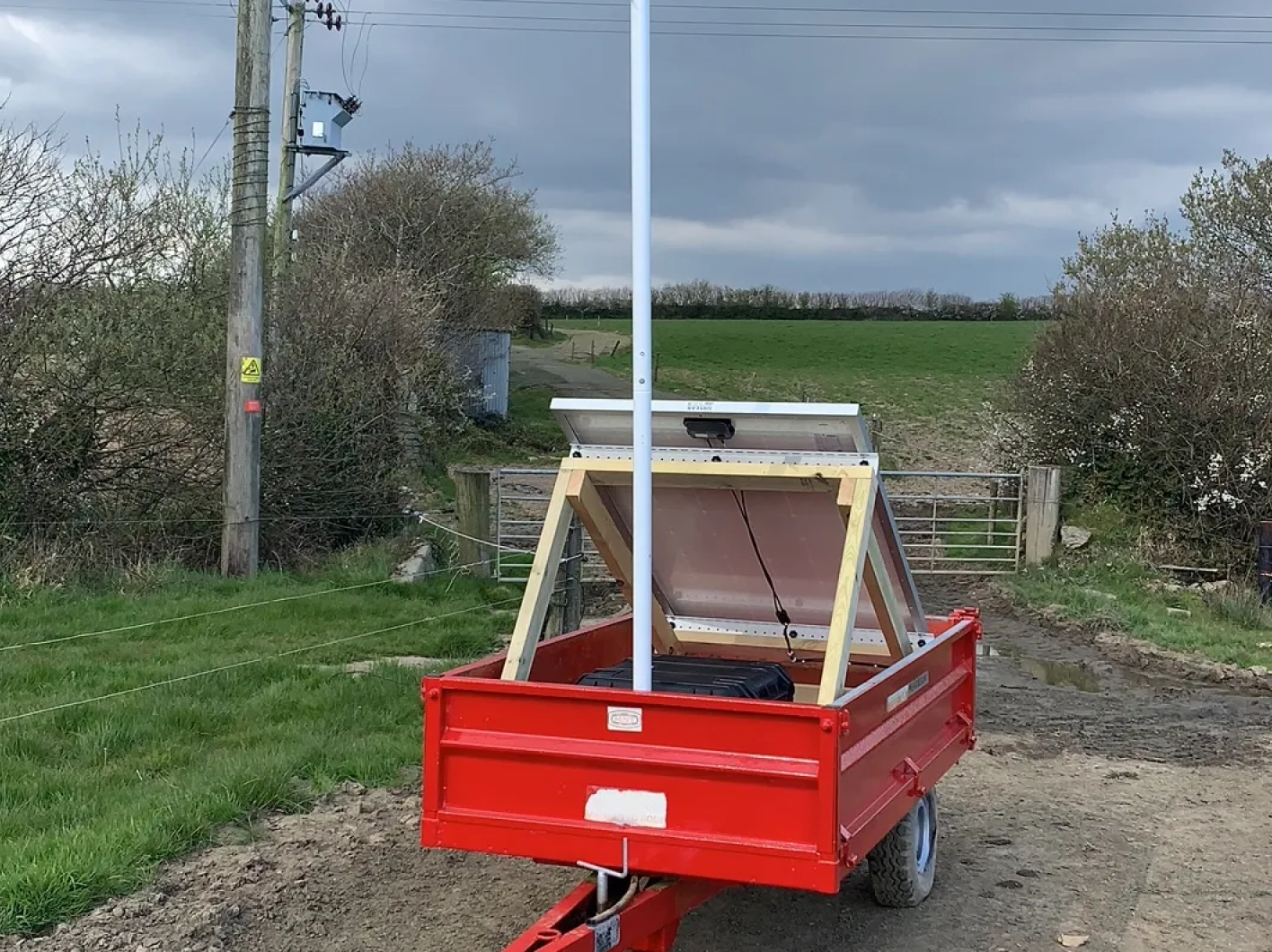 A red farm trailer carrying a solar panel array on a wooden frame with a tall white antenna mast, positioned on a farm track overlooking green fields.