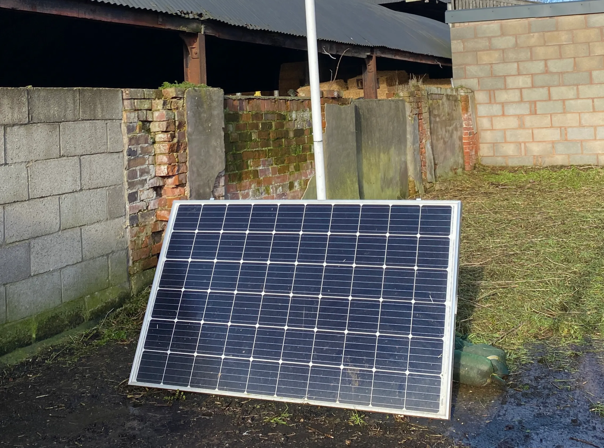 A single solar panel tilted toward the sun on a grassy area next to a farm building, used to provide renewable energy for agricultural equipment.
