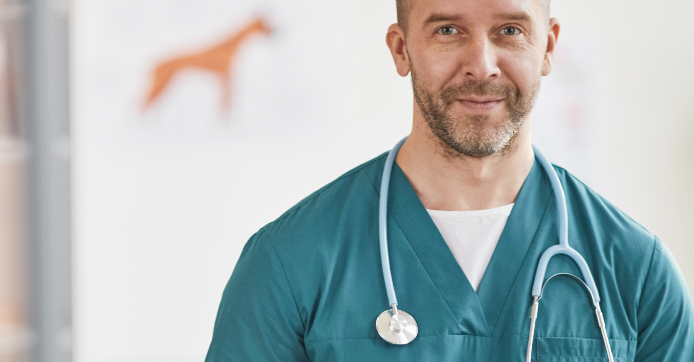 A smiling male veterinarian with a beard wearing teal scrubs and a blue stethoscope around his neck in a clinic setting.